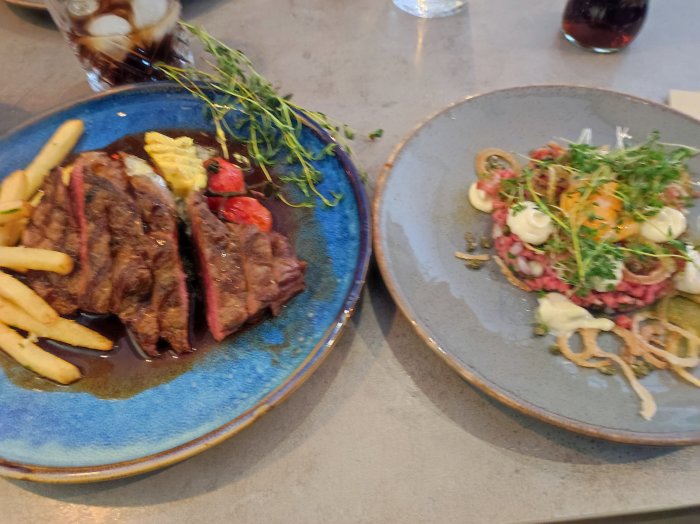 Två tallrikar med mat på en restaurang: grillad biff med pommes frites och en råbiff med äggula och kryddgrönt.