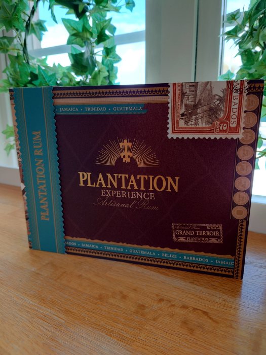 Flasklåda för Plantation Experience Artisanal Rum med frimärke, omgiven av gröna växter och fönster.