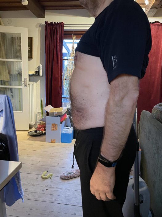Person i hemmiljö med upplyft tröja som visar magen, inrett rum i bakgrunden.