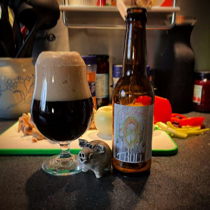 Ölstiligt glas fyllt med mörk öl bredvid en flaska med etiketten "TROLL", på en köksbänk med köksredskap i bakgrunden.