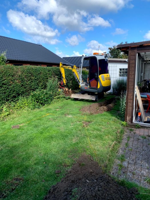 Minigrävare i trädgård gräver för altanbygge, med gräshoppar och jordhögar på gräsmatta.