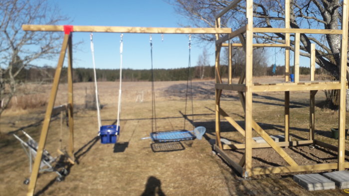 Nybyggd lekstuga med intilliggande gungställning på en gräsmatta, gungorna snett hängande.