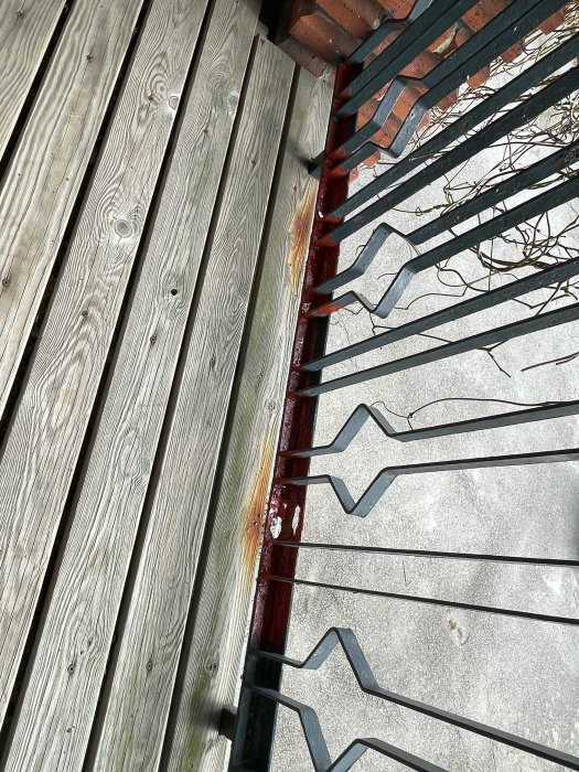 Närbild på balkongräcke med rostskador vid nederkanten intill träplankor.