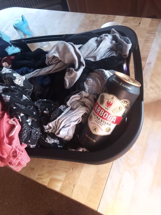 Burk med Arboga öl i en korg med blandade kläder på ett trägolv.