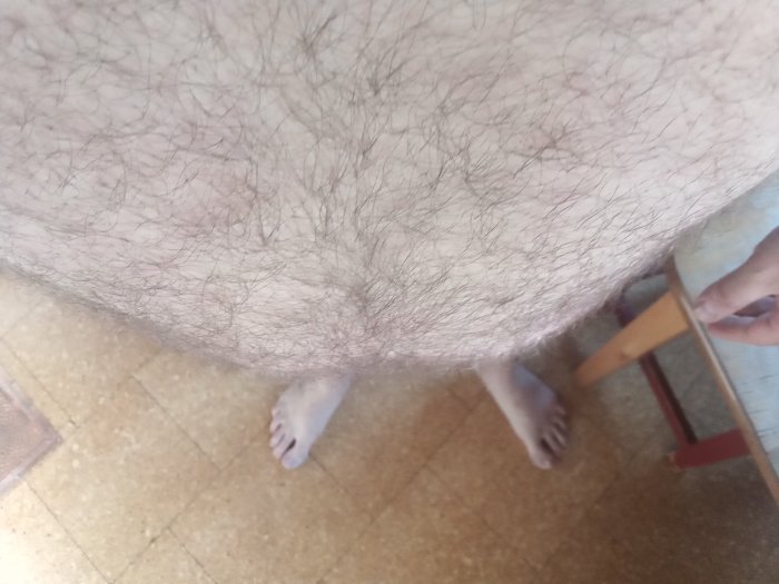 Nedifrån och upp-bild av en persons håriga ben som sitter med fötterna på ett golv och en stol syns delvis.