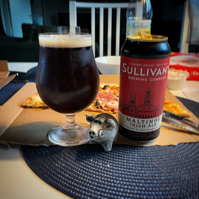Mörk ale i glas med Sullivan's flasketikett, grishuvudsparbössa framför, pizza i bakgrunden.
