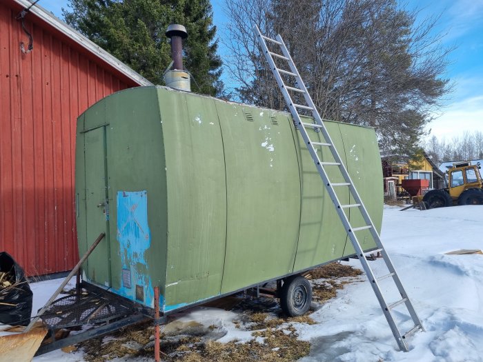 Sliten grön vagn med rundad vägg och kaminrör på sidan, står med en stege lutad mot den på snöklädd mark.