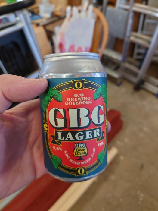 En hand håller en burk GBG Lager mot en bakgrund av en rörig verkstad.