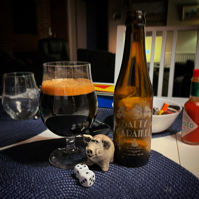 Ölglas fyllt med stout, en flaska Salted Caramel Stout, tärningar och en spargris på ett bord.