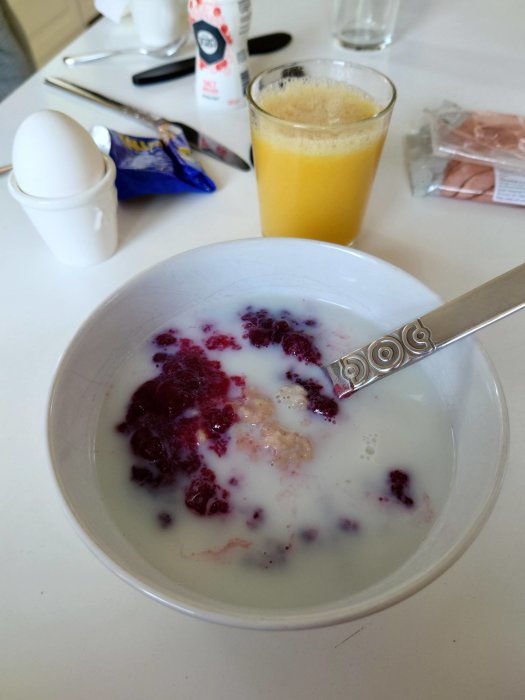 Frukost med havregrynsgröt och lingonsylt, ägg, juice, och skinkförpackning i bakgrunden.