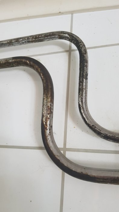 Kromade kopparrör med svart utfällning mot en bakgrund av vita kakelplattor.
