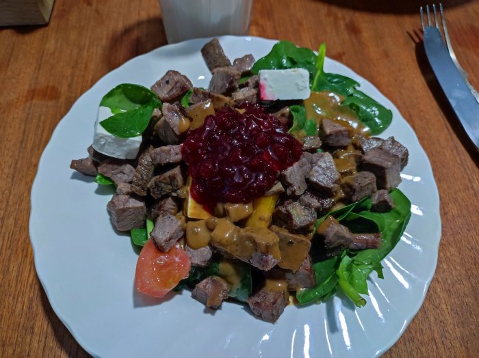 Tallrik med rågad portion av blandad sallad med tärnat kött, kuber av vit ost och lingonsylt, serverad med silverbestick.