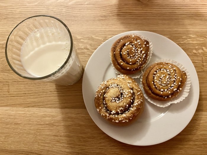 Ett glas mjölk och tre nybakade kanelbullar med pärlsocker på en vit tallrik, serverade på ett träbord.