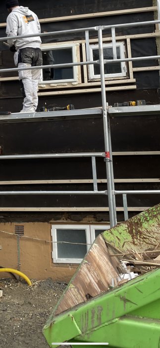 Arbetare monterar fasad med svarta plankor ovanför halvvisade källarfönster vid byggnation.