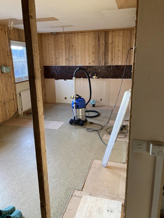 Renoveringsrum med halvavrivna väggbeklädnader, dammsugare och byggmaterial på golvet.
