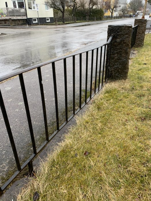 Snett svart smidesstaket med rostfläckar fastsatt vid stora stenpelare längs en gräsmatta.