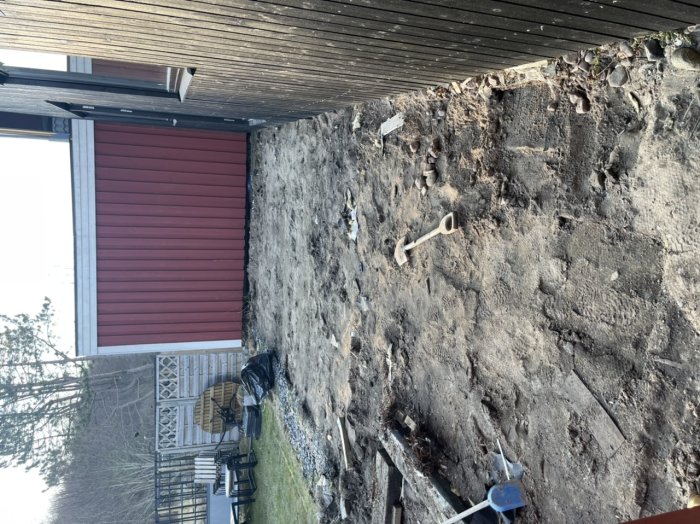 Upprivet markområde för byggprojekt nära rött hus med verktyg på marken.