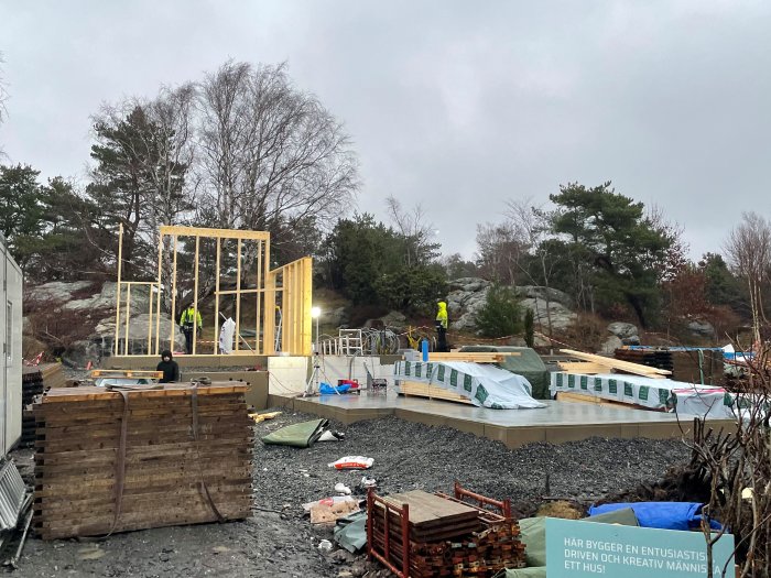 Byggplats med pågående arbete på en framtida entré med högt i tak och genomsikt, omgivet av byggmaterial och natur.