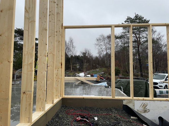 Byggnadsstomme i trä med höga pelare och en framtida genomskinlig entré under uppbyggnad, omgiven av byggmaterial.