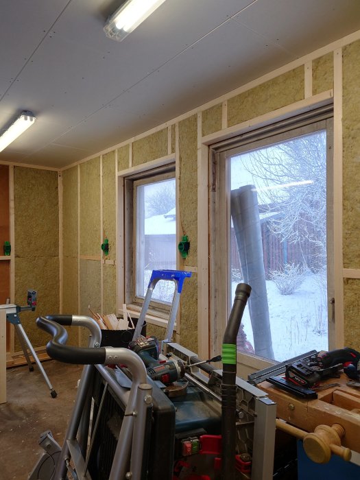 Inomhusbyggarbetsplats med ny isolering på väggen, verktyg framför och utsikt mot snöig natur genom fönstret.