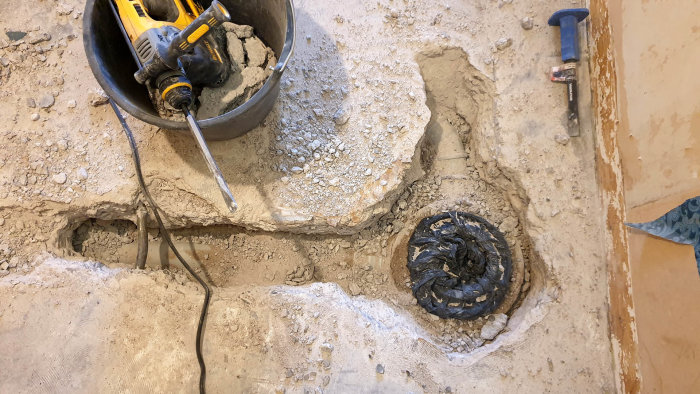 Uppbruten betonggolv med synliga rör, verktyg, och gammal skadad avloppsinstallation.