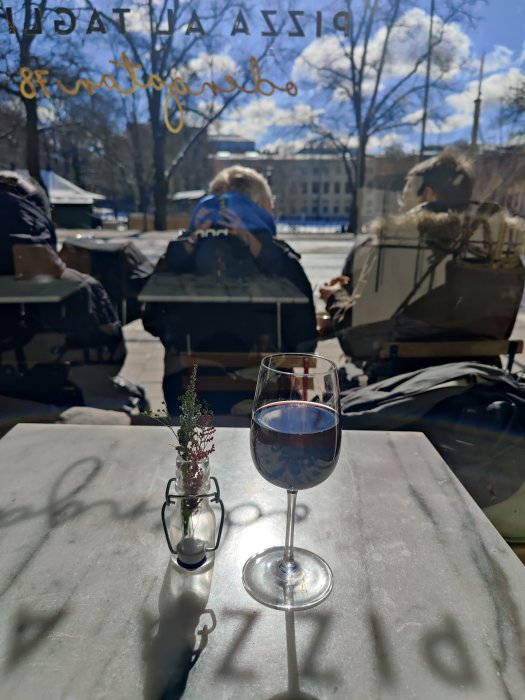 Ett glas vin och en blomvas på uteserveringsbord med personer och soligt vårväder i bakgrunden.