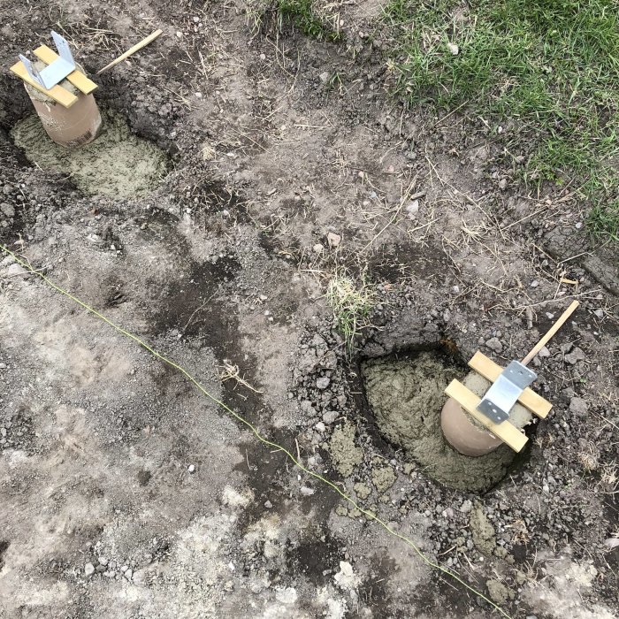 Två betongfundament med papprör och balksko förberedda för stolpar i en grävd mark.