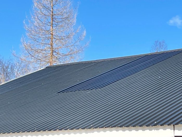 Vågformad metalltakbeklädnad med några mindre skador mot en klarblå himmel och träd i bakgrunden.