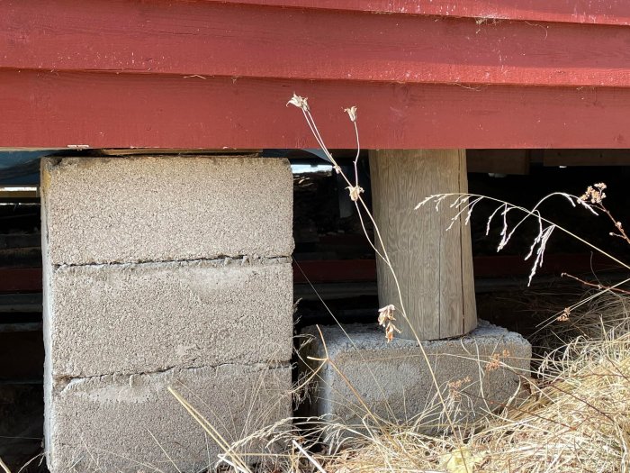 Grundpelare av hus med sned stolpe och lossande plastmatta under rödmålad stuga.