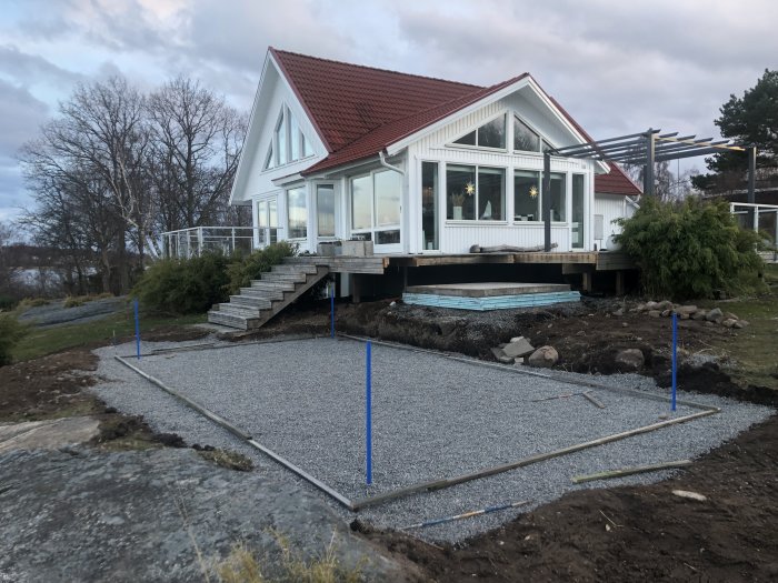 Grund för poolbygge med grus och markeringsstolpar framför ett hus, arbete pågår.