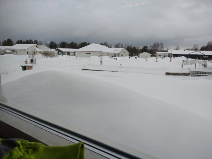 Utsikt över snötäckt bostadsområde med hus och trädgårdar sett från ett fönster.