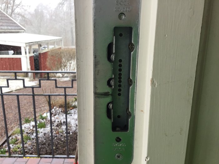 Närbild på en öppen ytterdörrs gångjärnssida med utsikt mot ett staket och snöfläckad trädgård.