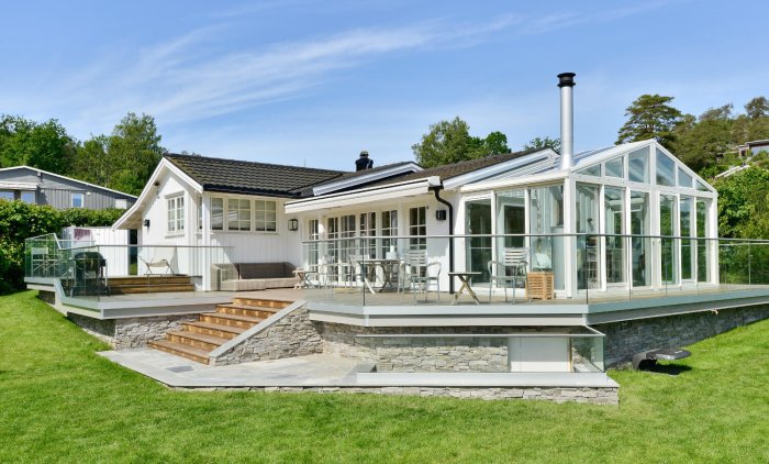 Vit villa med uterum i glas, stentrappa och terrass med glasräcke på grön gräsmatta.