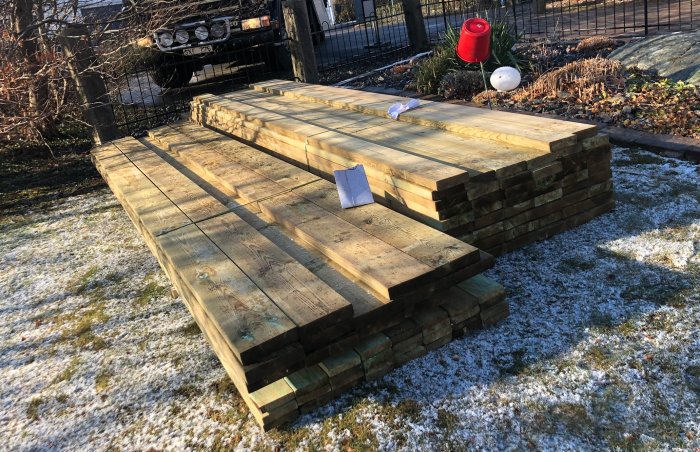 Virke staplat på gräsmark med snöfläckar för projektet att renovera en brygga.