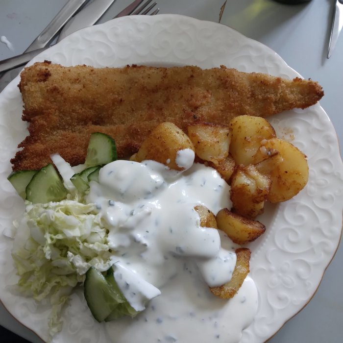 Panerad fisk med citron- och gräslöksmajonnäs, stekt potatis och sallad på en tallrik.