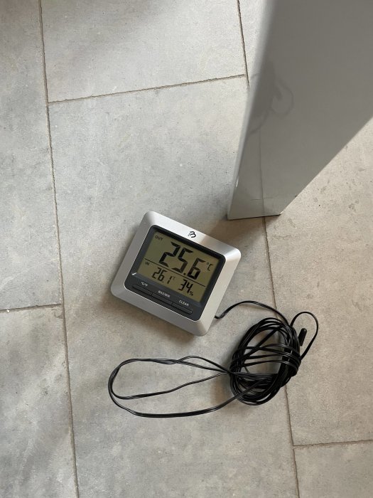 Digital termometer med display visande inomhus- och utomhustemperatur placerad på ett kakelgolv.