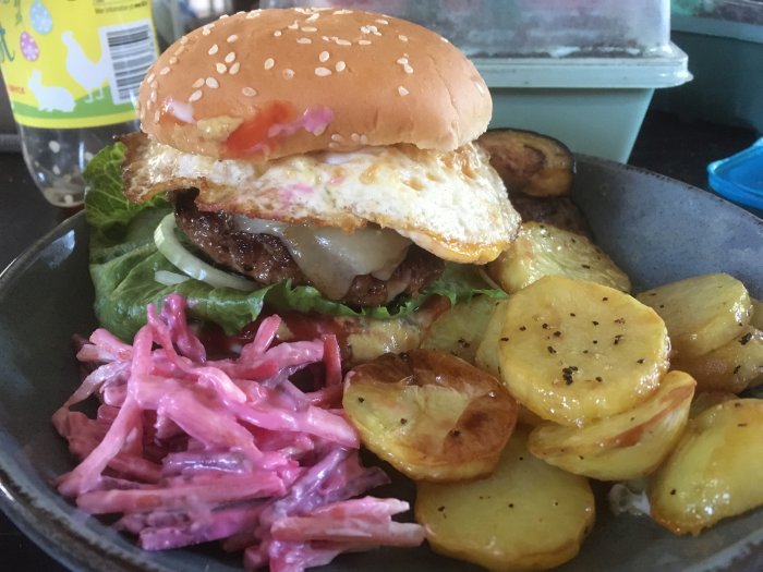 Hemgjord hamburgare med ägg och råa ingredienser på ett sesambröd, coleslaw och ugnsstekta potatisskivor.