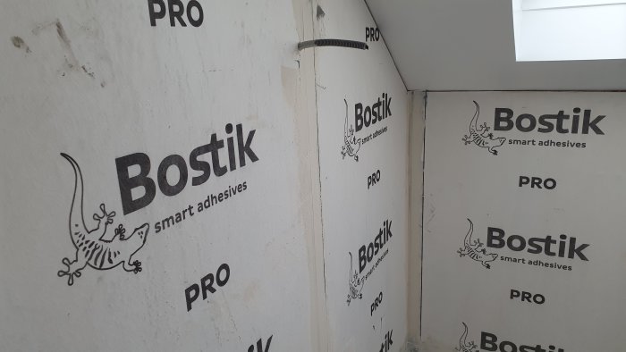 Hörn av ett rum med isolerade väggar täckta av Bostik smart adhesives-logotyper och en öppen kabelkanal.