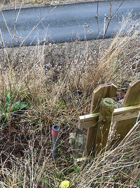 Gränsrör vid ett fritidshus, dokumenterat bland vissna gräs och ett trästaket, med väg i bakgrunden.