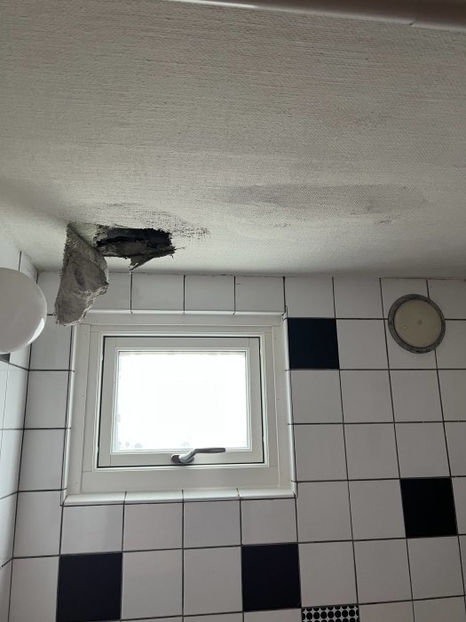 Skadat badrumstak med hål nära fönster, vita och svarta kakelplattor på väggen.