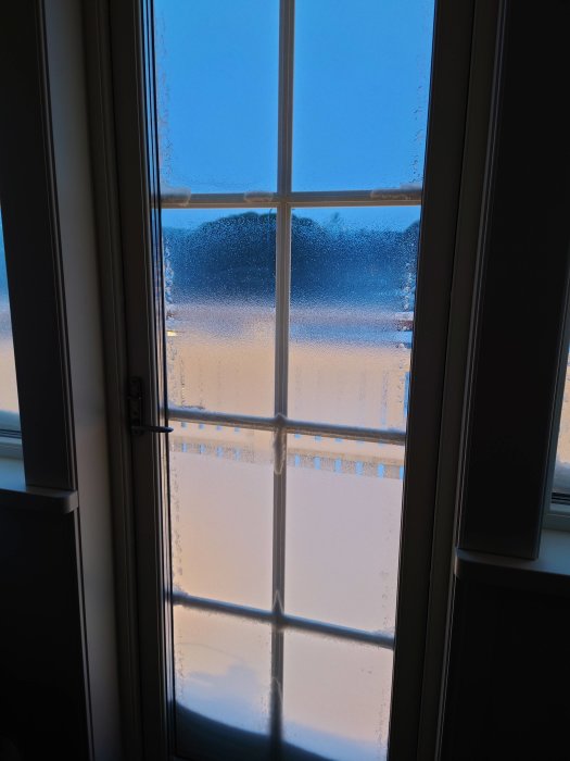 Fönster med kondens och isbildning på insidan, som delvis skymmer utsikten mot en blå himmel.