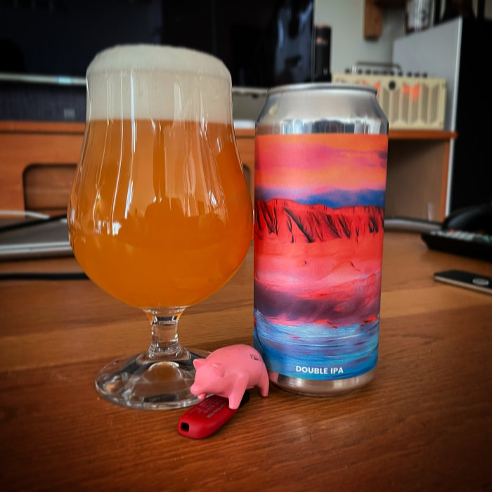 Ölglas fyllt med grumlig öl bredvid en burk Double IPA och en liten rosa plastgris på ett bord.