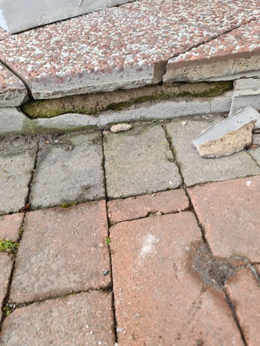 Sprickor i husfasad vid stenplattor med en lös bit av tegelmurverk nära marken.