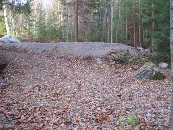 En skogstomt med en hög av grus och stenar, kapade träd i bakgrunden, och en grusad markyta framför.