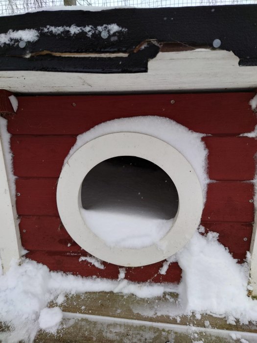 Röd byggnad med snöblockerad rund öppning, skadat svart tak och vit snö.