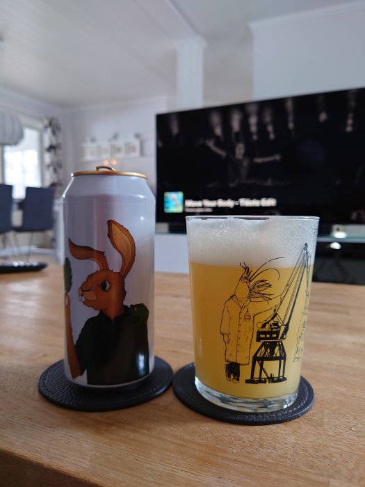 Ölburk och glas med konstnärliga etiketter, "Hoppy Friends" från Fermenterarna, på ett bord framför en TV.