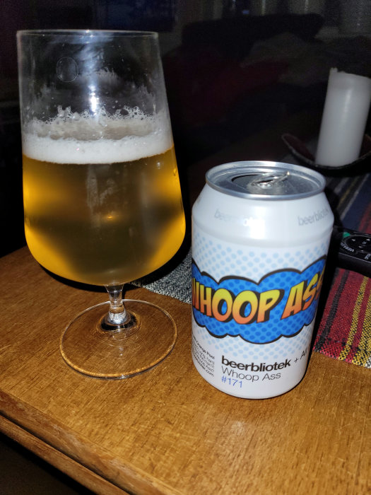 Glas med öl bredvid en öppnad ölburk med färgstark etikett på ett träbord.