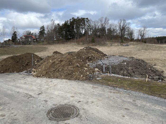 Jordhögar och schaktmassor vid en byggarbetsplats nära en väg, med nivåjusteringar under arbete.