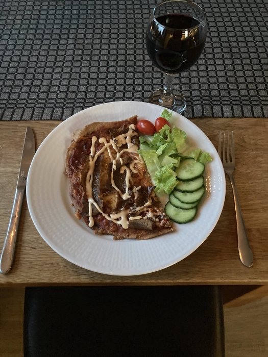 Kebabpizza på tallrik med sallad och tomater bredvid ett glas rödvin på dukat bord.