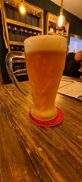 En stor skummande öl i sejdel på en röd ölbricka, interiör av bar i bakgrunden.
